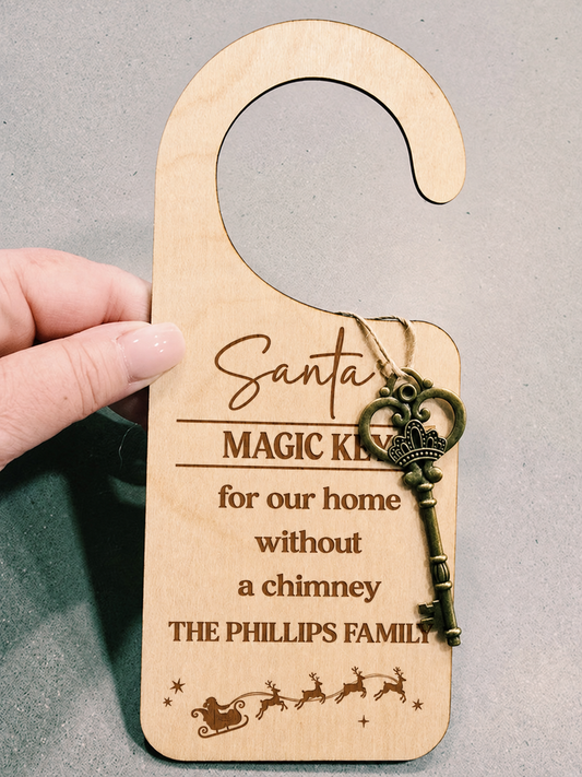 A hand holds a personalized wooden door hanger key tag engraved with 'Santa's Magic Key for our home without a chimney THE PHILLIPS FAMILY' and decorated with an antique-style key charm, against a neutral gray background.