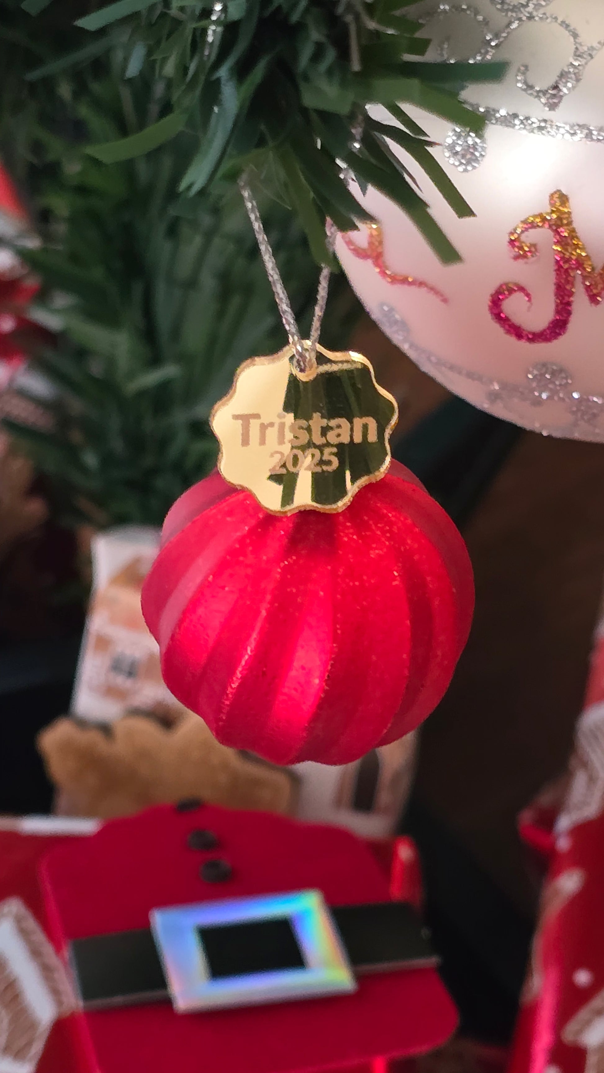 Red ornament with a gold tag labeled 'Tristan' on a Christmas tree.
