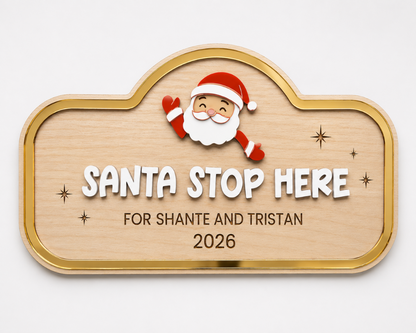A personalized wooden plaque with a gold border, featuring a cartoon Santa Claus illustration above the text 'SANTA STOP HERE FOR SHANTE AND TRISTAN 2026' on a light gray background.