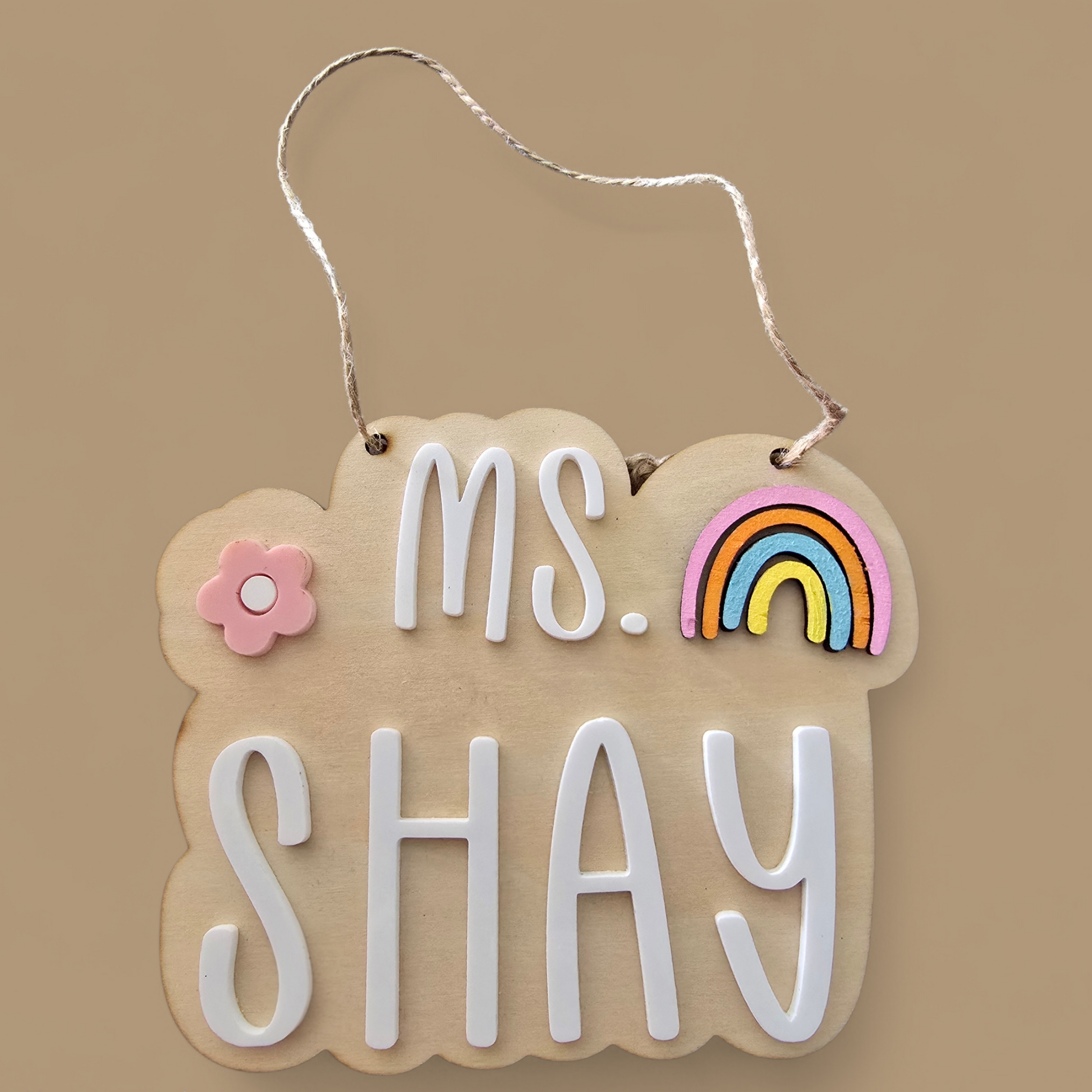 Decorative sign with 'ms. SHAY' text, rainbow, and flower on a beige background