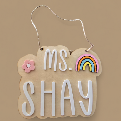 Decorative sign with 'ms. SHAY' text, rainbow, and flower on a beige background
