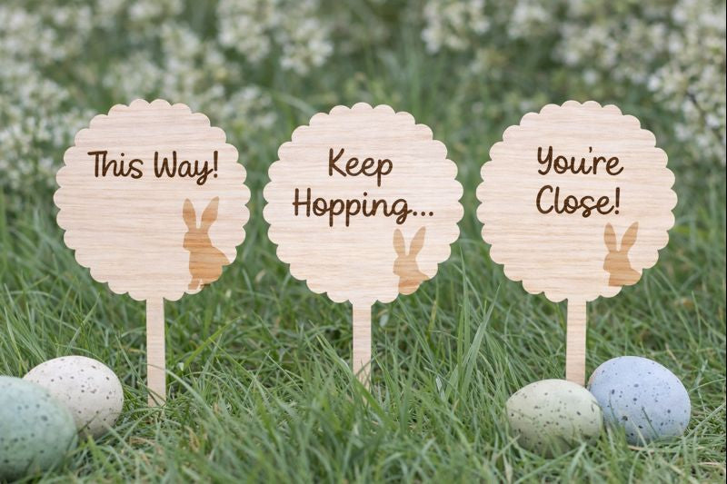 Wooden signs with bunny illustrations and text on sticks in grass with Easter eggs.