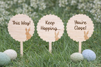 Wooden signs with bunny illustrations and text on sticks in grass with Easter eggs.