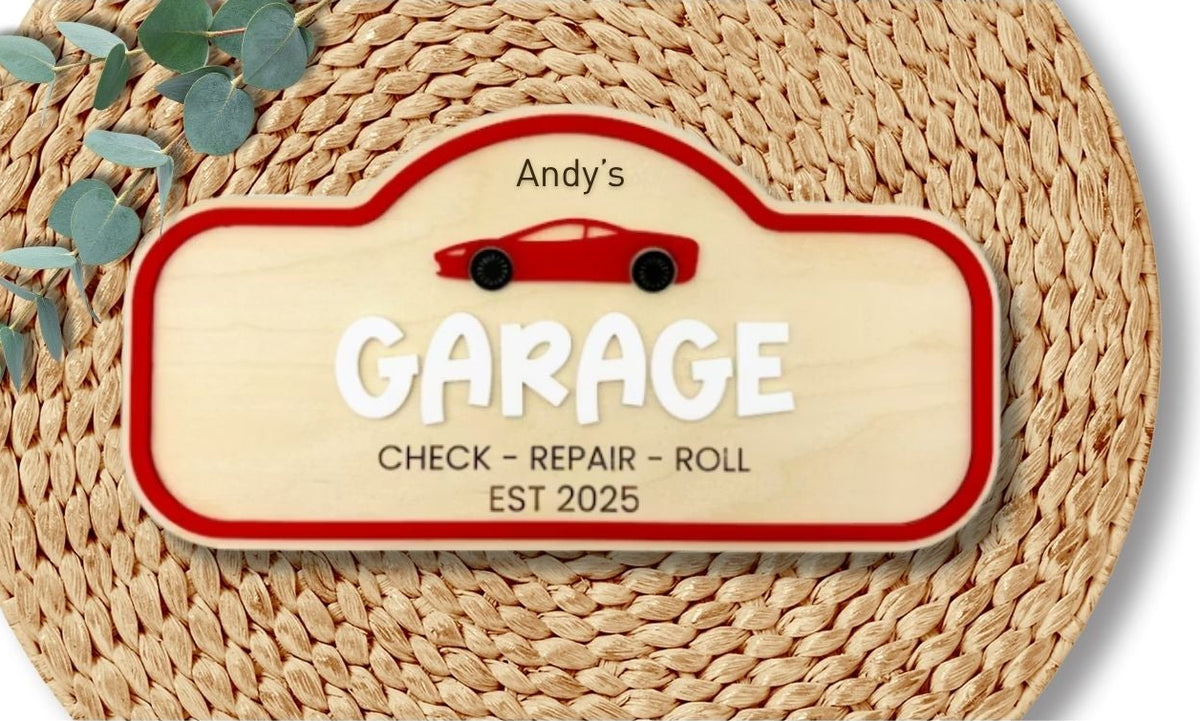 A personalized wooden garage sign with a red border and white lettering that reads 'Andy's GARAGE CHECK - REPAIR - ROLL EST 2025', placed on a woven straw mat with green leaves in the corner, shown on a white background.