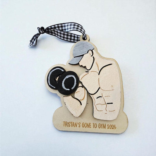 Wooden ornament with a man lifting weights and text 'Tristan's Gone to Gym 2025' on a light blue background.