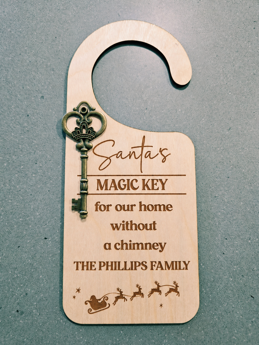 A personalized wooden Christmas door hanger key for 'Santa's Magic Key' with an engraved key and sledding graphic, shown on a textured gray background.