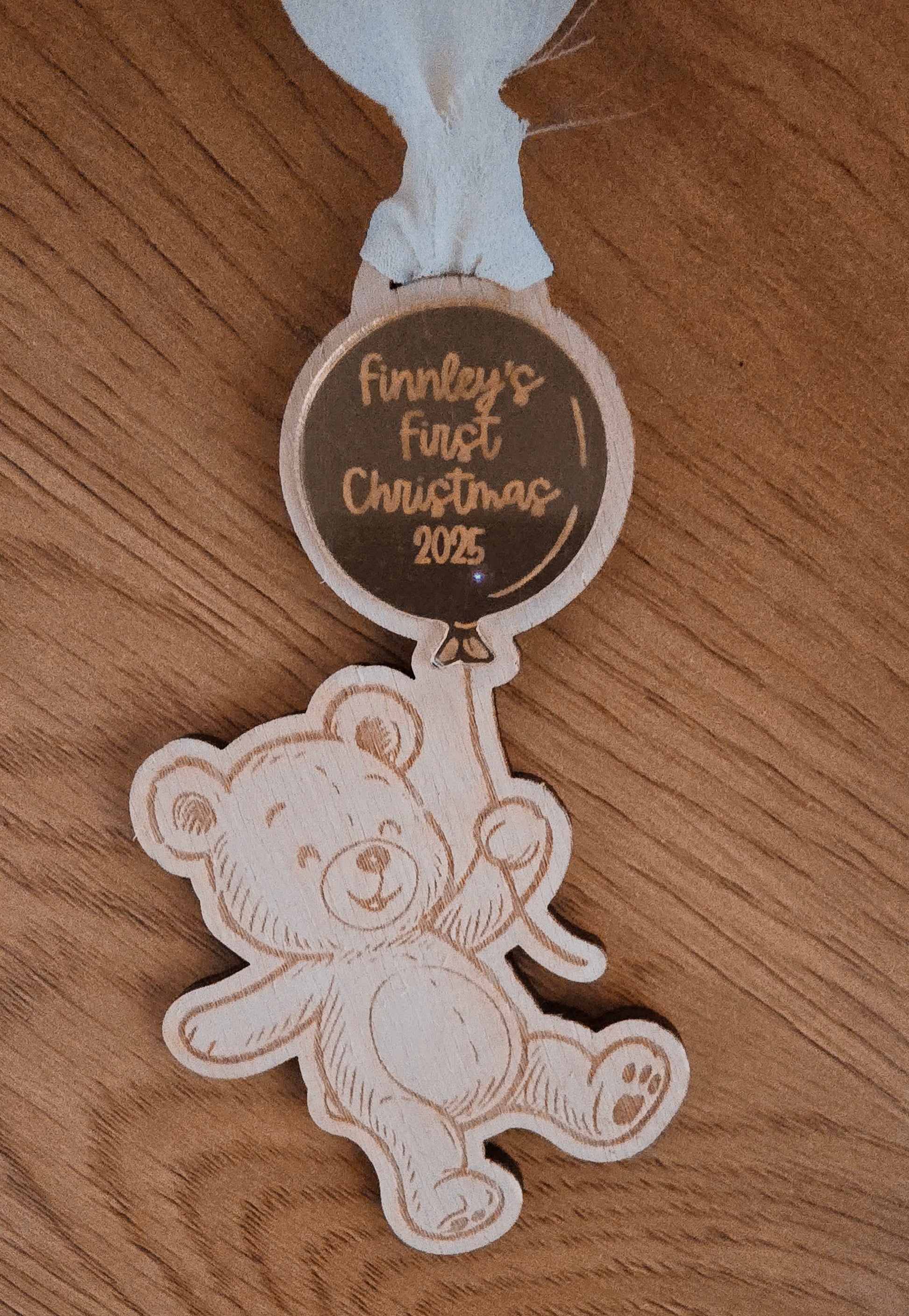 Decorative tag with a teddy bear design and 'Finnley's First Christmas 2023' text on a wooden surface.
